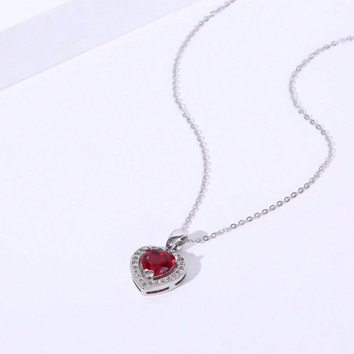 Collar con dije de corazón en color plateado, con una piedra central rojo y borde de circonias.