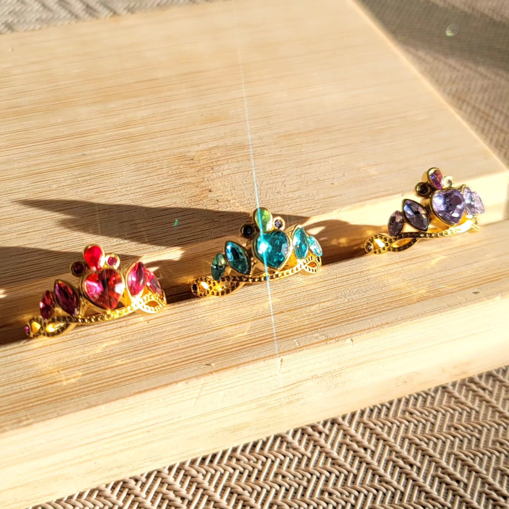 Anillos de princesas Disney, cada uno con un dije en forma de corona en diferentes colores: fucsia, blanco y morado.

