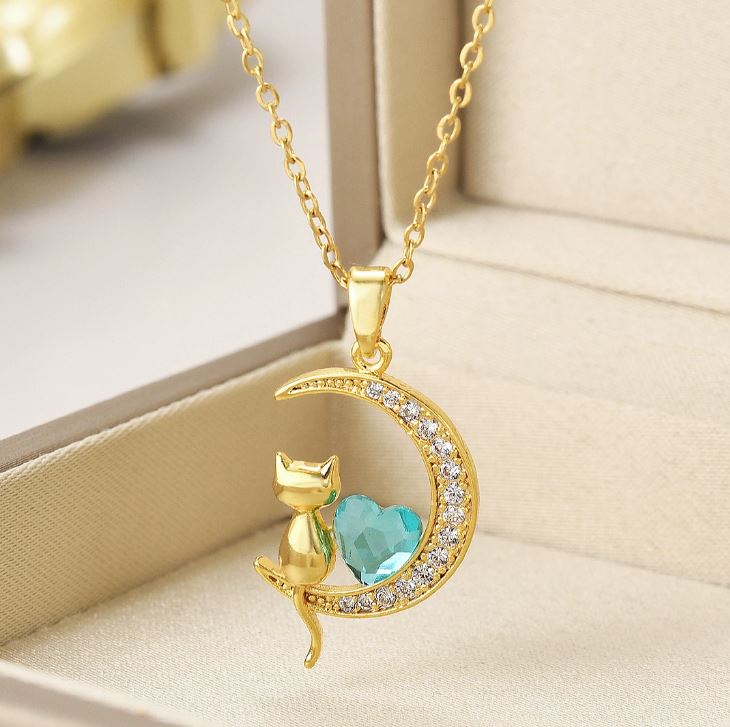 Collar con dije de gatito sentado en una luna, con un corazón central de cristal azul y acabado dorado.

